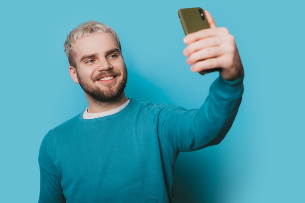 Photo monochrome d'un homme de race blanche aux cheveux blonds et barbe faisant un selfie à l'aide d'un téléphone sur un mur bleu