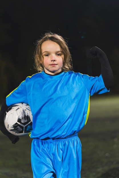 Une photo moyenne d'une petite fille mignonne en uniforme bleu tenant un ballon de football et montrant des enfants puissants