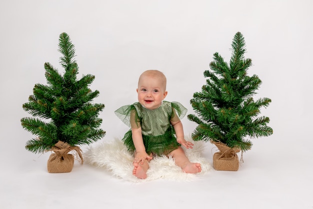 Photo de Noël une petite fille enfant assise près des arbres de Noël dans une robe verte sur blanc