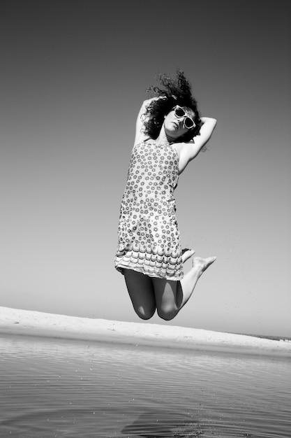 Photo en noir et blanc d'une femme aux cheveux noirs bouclés sautant dans une robe de soleil courte