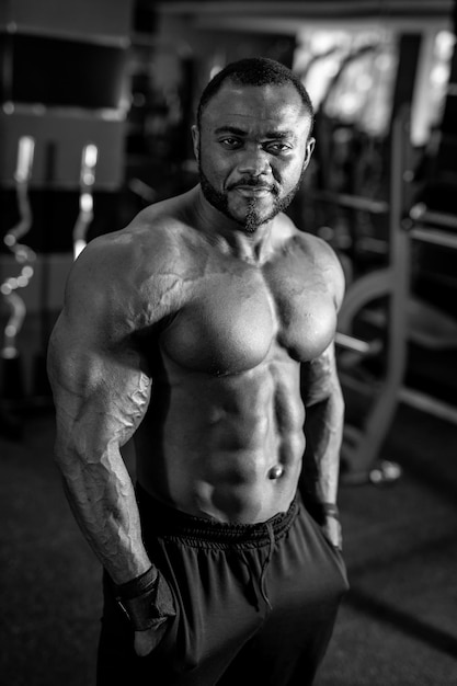 Photo en noir et blanc de l'homme fort avec un type de corps musclé posant dans une salle de sport moderne Homme multiracial regardant la caméra avec un léger sourire