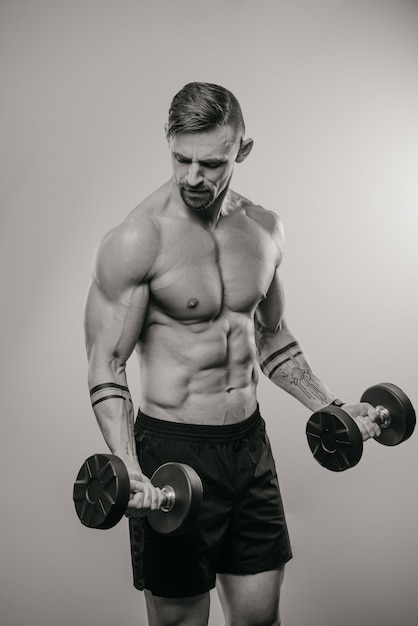 Une photo en noir et blanc d'un homme musclé avec une barbe qui fait des boucles de biceps avec des haltères