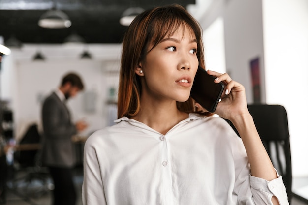 Photo de la pensée d'une femme d'affaires asiatique regardant de côté et parlant au téléphone portable dans un bureau à aire ouverte