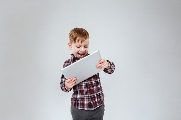 Photo d'un petit enfant mignon debout sur un mur gris tenant une tablette. Regardez la tablette.