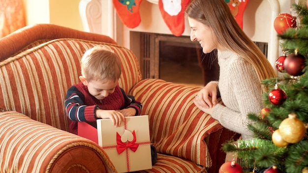 La photo d'un petit garçon souriant et heureux ouvre une boîte avec un cadeau de Noël et regarde à l'intérieur. Mère donnant un cadeau à son enfant le nouvel an