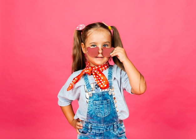 Photo d'une petite belle fille mignonne debout isolée sur fond rose, portant des lunettes de soleil, regardant la caméra.