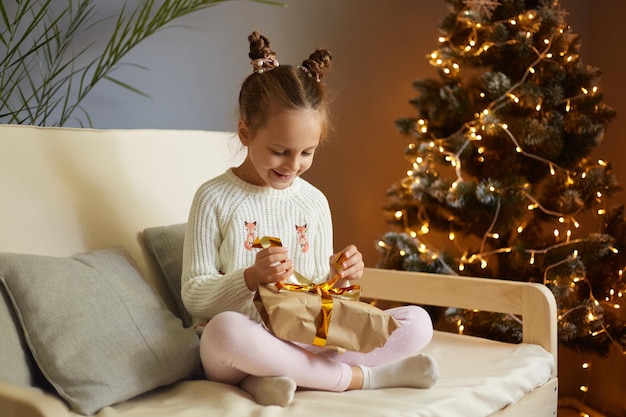 Photo d'une petite fille assez heureuse et satisfaite avec deux chignons portant des vêtements décontractés assis sur un canapé et ouvrant une boîte cadeau pour Noël regardant souriant au cadeau