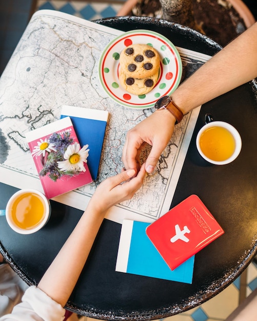 Photo à plat de mains d'homme et de femme se touchant. Paire de planification de vacances. Sur la table noire différents guides de voyage, carte, tasses à thé, biscuits et passeport. Notion d'envie d'errance.