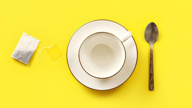 Photo à plat, sachet de thé, tasse et cuillère en porcelaine vide sur tableau jaune.