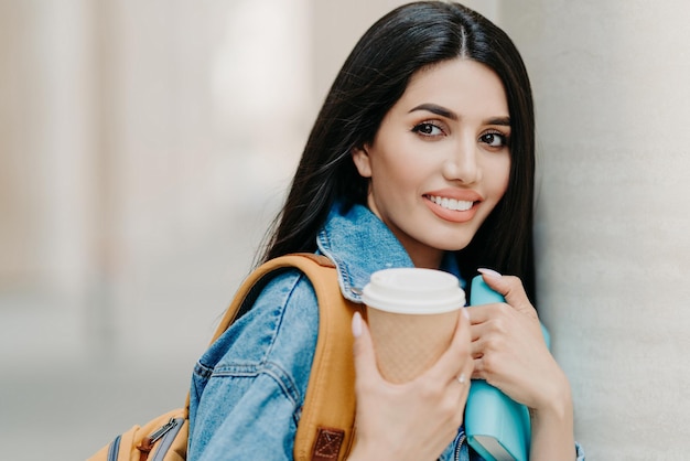 Photo en plein air d'une étudiante brune maquillée, cheveux longs noirs, vêtue d'une veste en jean, tient un café à emporter, un manuel, porte un sac à dos, regarde quelque part au loin, se promène dans la ville