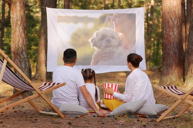 Photo en plein air d'une famille positive assise dans la forêt sur des chaises longues avec leur petite fille et regardant un film sur un projecteur regardant un film sur un chiot ou regardant des photos