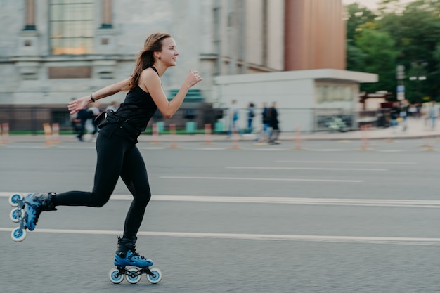 Photo en plein air d'une femme aux cheveux noirs active en bonne forme physique qui aime les sports extrêmes sur des rouleaux le long de la route dans une rue animée se détend pendant le week-end en plein air. Concept de mode de vie sain