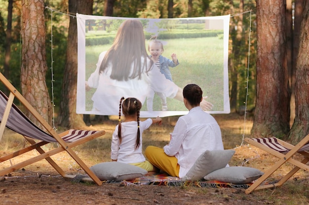 Photo en plein air d'une mère et d'une petite fille assise sur le sol sur une couverture et regardant une photo sur un projecteur profitant de passer du temps en plein air dans une belle forêt de pins