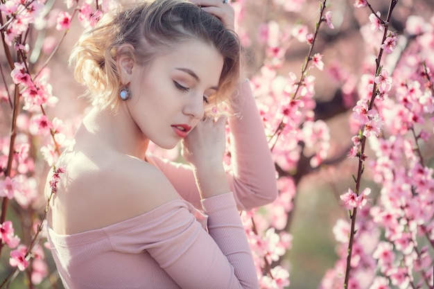 Photo en plein air de mode de la magnifique jeune femme en robe élégante posant dans le jardin avec des pêchers en fleurs. Blonde dans les jardins fleuris