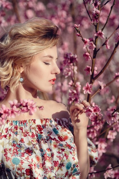 Photo en plein air de mode de la magnifique jeune femme en robe élégante posant dans le jardin avec des pêchers en fleurs. Blonde dans les jardins fleuris