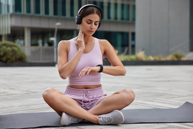 Une photo en plein air d'une sportive vérifie le pouls après l'entraînement garde la main sur le cou regarde la montre-bracelet assise les jambes croisées sur un karemat vêtu de trains de vêtements de sport en plein air