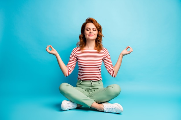 Photo pleine grandeur d'une femme calme et sereine assis plancher croisé les jambes montrent om signe exercice yoga méditation porter élégant tenue verte isolée sur la couleur bleue