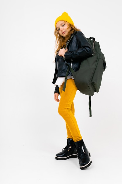 Photo pleine longueur d'une adolescente élégante dans une chemise blanche, une veste en cuir noire déboutonnée, un pantalon jaune et un chapeau jaune, avec un sac à dos sur les épaules posant sur blanc