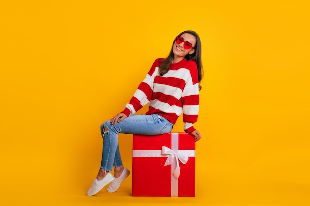 Photo pleine longueur d'une belle femme brune élégante et excitée dans des lunettes de soleil alors qu'elle est assise et pose avec une grande boîte-cadeau rouge