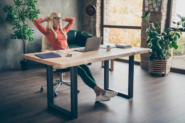 Photo pleine longueur de calme positif femme pacifique propriétaire de l'entreprise mandataire finir le travail de démarrage s'asseoir table se détendre fermer les yeux étirer les mains dans bureau col roulé rouge loft