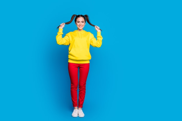 Photo pleine longueur d'une dame drôle et joyeuse tenir les bras à deux queues bonne humeur usure ludique pull à capuche jaune décontracté pantalon rouge baskets isolé fond de couleur bleu vif