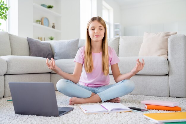 Photo pleine longueur d'une enfant focalisée étudie à distance un tapis de sol assis les jambes croisées les yeux fermés méditent l'équilibre de la pratique avant la leçon d'apprentissage en ligne dans la maison à l'intérieur