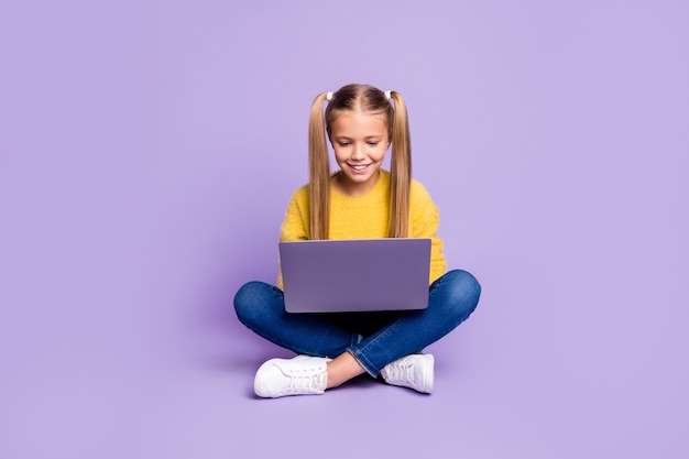 Photo pleine longueur de l'enfant intéressé positif assis les jambes croisées travail plié son ordinateur portable lire les médias sociaux nouvelles portent des vêtements de style décontracté isolé sur mur de couleur violet