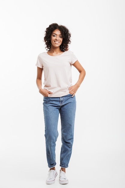 Photo pleine longueur de femme américaine vivace portant des jeans et t-shirt posant avec un sourire candide et les mains dans la poche, isolé sur un mur blanc