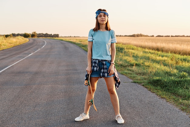 Photo pleine longueur d'une femme confiante et satisfaite portant un short, un t-shirt, un bandeau pour les cheveux, tenant une planche à roulettes dans les mains, regardant la caméra, après avoir fait de la planche à roulettes en plein air.