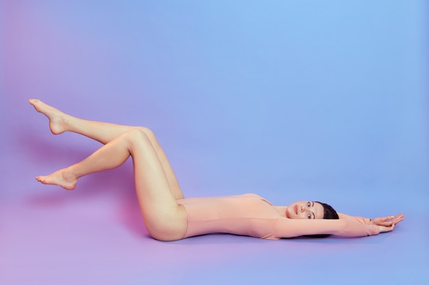 Photo pleine longueur de femme avec un corps parfait, allongée sur le sol et étirant les bras, levant de longues jambes, habille un body beige, isolée sur un mur bleu avec une lumière néon rose.