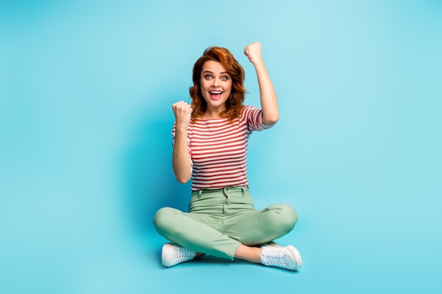 Photo pleine longueur d'une femme excitée s'asseoir les jambes croisées célébrer sa victoire de loterie triomphe lever les poings crier ouais porter des vêtements de bonne tenue isolés sur la couleur bleue