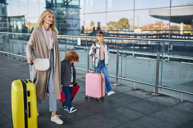 Photo pleine longueur d'une femme marchant avec deux enfants avec des bagages le long de la clôture en verre jusqu'à l'aéroport