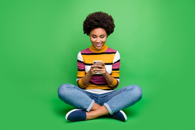 Photo pleine longueur fille afro-américaine joyeuse s'asseoir jambes pliées utiliser téléphone intelligent lire les nouvelles du réseau social commentaire suivre republier porter des jeans vêtements lumineux