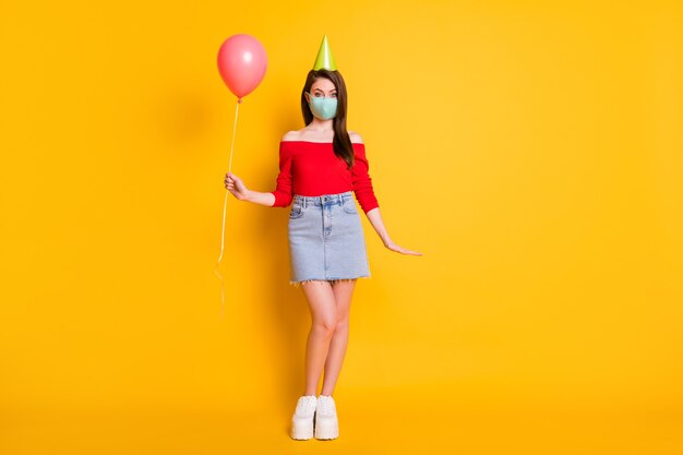 Photo pleine longueur d'une fille positive portant un masque médical a une fête d'anniversaire covid célébration de quarantaine tenir un ballon porter un jean haut rouge jupe jambes cône isolé fond de couleur brillant brillant