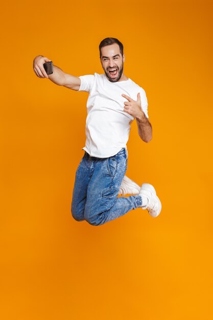 Photo pleine longueur de gars positif de 30 ans en tenue décontractée en riant et en prenant selfie sur téléphone portable, isolé