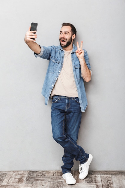 Photo pleine longueur de l'homme de race blanche brune ayant barbe prenant selfie sur téléphone portable et montrant la paix chanter isolé sur mur gris