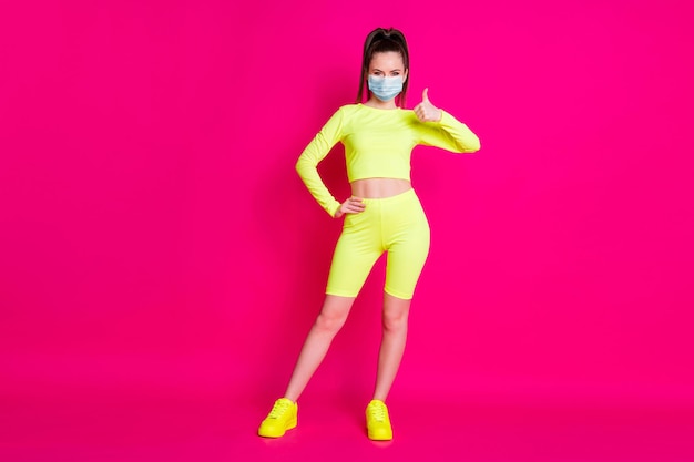 Photo pleine longueur de jeune femme habillée sportwear masque main pouce vers le haut bras taille isolé fond de couleur rose