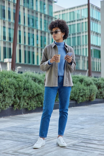 Photo pleine longueur d'une jeune femme à la mode porte une veste en jean, des baskets blanches et des lunettes de soleil tient un téléphone portable et boit des poses de café en milieu urbain. Concept de technologie de style de vie moderne.