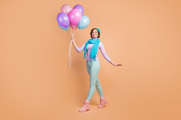 Photo pleine longueur d'une jolie dame portant de nombreux ballons à air marchant fête surprise meilleur ami porter un pull violet lilas pantalon vert bottes écharpe béret bleu isolé fond de couleur beige