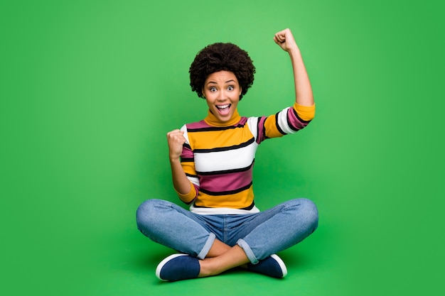 Photo pleine longueur de joyeuse fille afro-américaine chanceuse heureuse s'asseoir les jambes croisées pliées gagner la loterie lever les poings crier ouais porter une bonne tenue de look