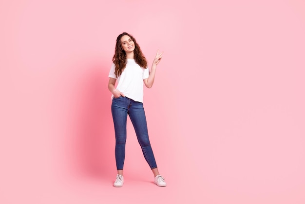 Photo pleine taille d'une jeune jolie fille de bonne humeur montre aux doigts la paix cool v-symbole isolé sur fond de couleur rose.