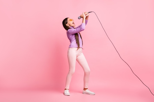 Photo pleine taille d'une petite fille chantant une chanson sur un micro portant un pantalon de chandail violet isolé sur fond de couleur pastel