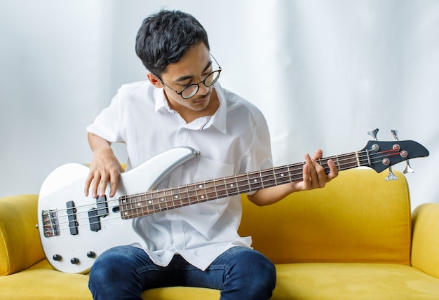 Photo de portrait d'un beau jeune adolescent souriant appréciant de jouer de la guitare basse. Guitariste junior en tenue décontractée assis sur le canapé jaune et tenant un instrument en regardant la caméra