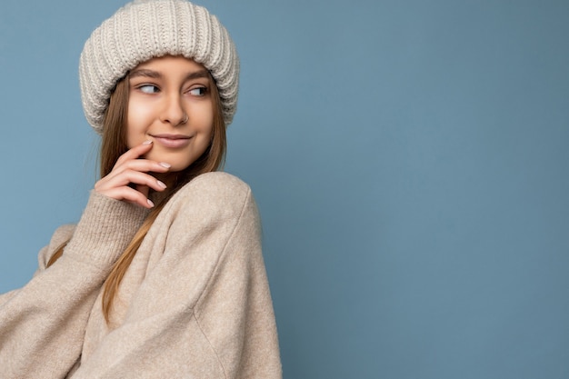 Photo photo de portrait de belle sexy souriante heureuse jeune femme blonde debout isolée sur un mur de fond bleu portant un pull beige et un chapeau beige regardant sur le côté et flirtant