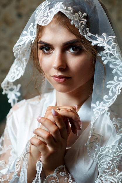 Photo portrait en gros plan d'une mariée maquillée professionnellement, elle est en blouse blanche, voile sur le visage. Jour de mariage de la jeune femme. Séance photo, coiffure, beauté