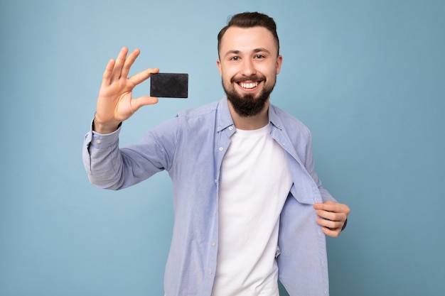 Photo prise de beau jeune homme barbu brune souriante portant une chemise bleue élégante et un t-shirt blanc isolé sur un mur de fond bleu tenant une carte de crédit en regardant la caméra