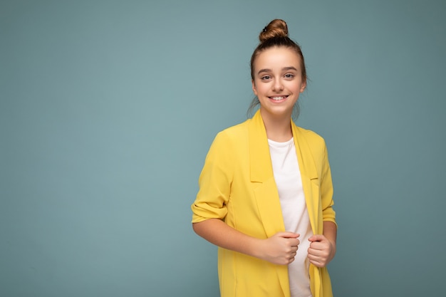 Photo prise de belle petite fille brune souriante et heureuse portant une veste jaune à la mode et un t-shirt blanc debout isolé sur un mur de fond bleu regardant la caméra