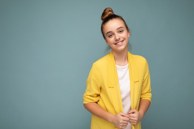 Photo prise de belle petite fille brune souriante et positive portant une veste jaune à la mode et un t-shirt blanc debout isolé sur un mur de fond bleu regardant la caméra