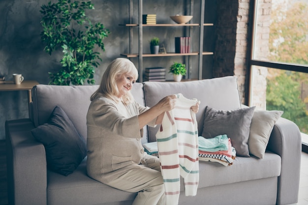 Photo de profil d'étonnante mamie âgée aux cheveux blancs tenant des mains pull bien rangées le mettre dans la pile de nettoyage général de printemps maison assis divan salon à l'intérieur