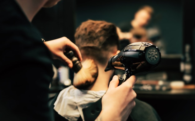 Photo de profil d'un jeune coiffeur coupant les cheveux de son client avec un rasoir électrique et un peigne dans un salon de coiffure.